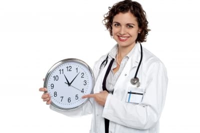 Female doctor holding a clock, pointing to it, emphasizing the importance of time management in pediatric board exam preparation.