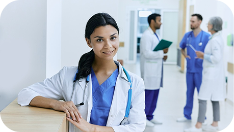 Smiling female doctor in a hospital hallway with a stethoscope around her neck in focus foreground while a team of medical professionals is in discussion in the background