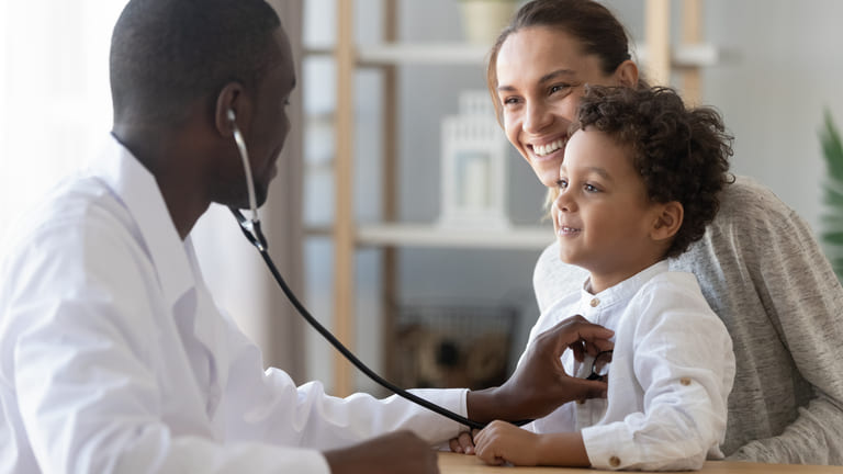A pediatrician using a stethoscope to examine a young child, while the child’s mother watches with a smile.