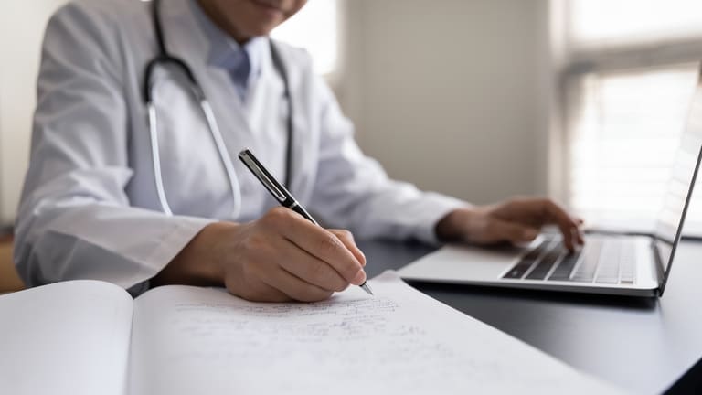 A doctor in white coat writing notes on a notepad while using a laptop on the desk.