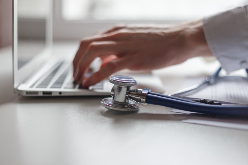 Close-up of a stethoscope on a desk, with a doctor's hand typing on a laptop keyboard in the background.