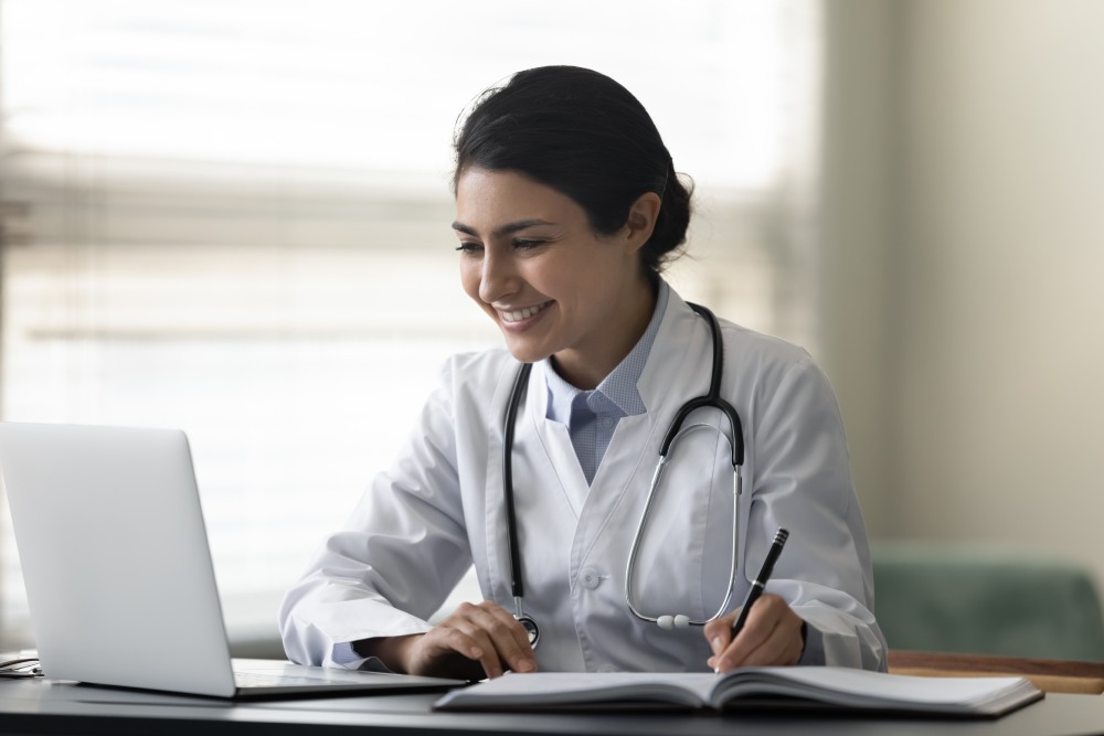 A female doctor smiling while working on a laptop, taking notes in a notepad at the same time.