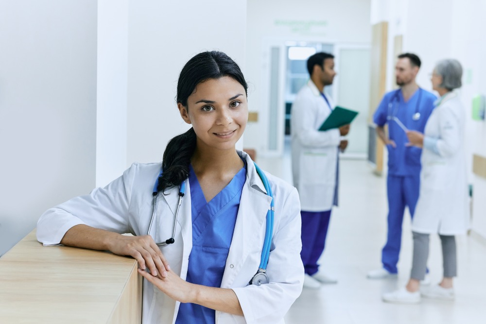 A female doctor standing confidently with other medical professionals in the background, all wearing scrubs and white coats.